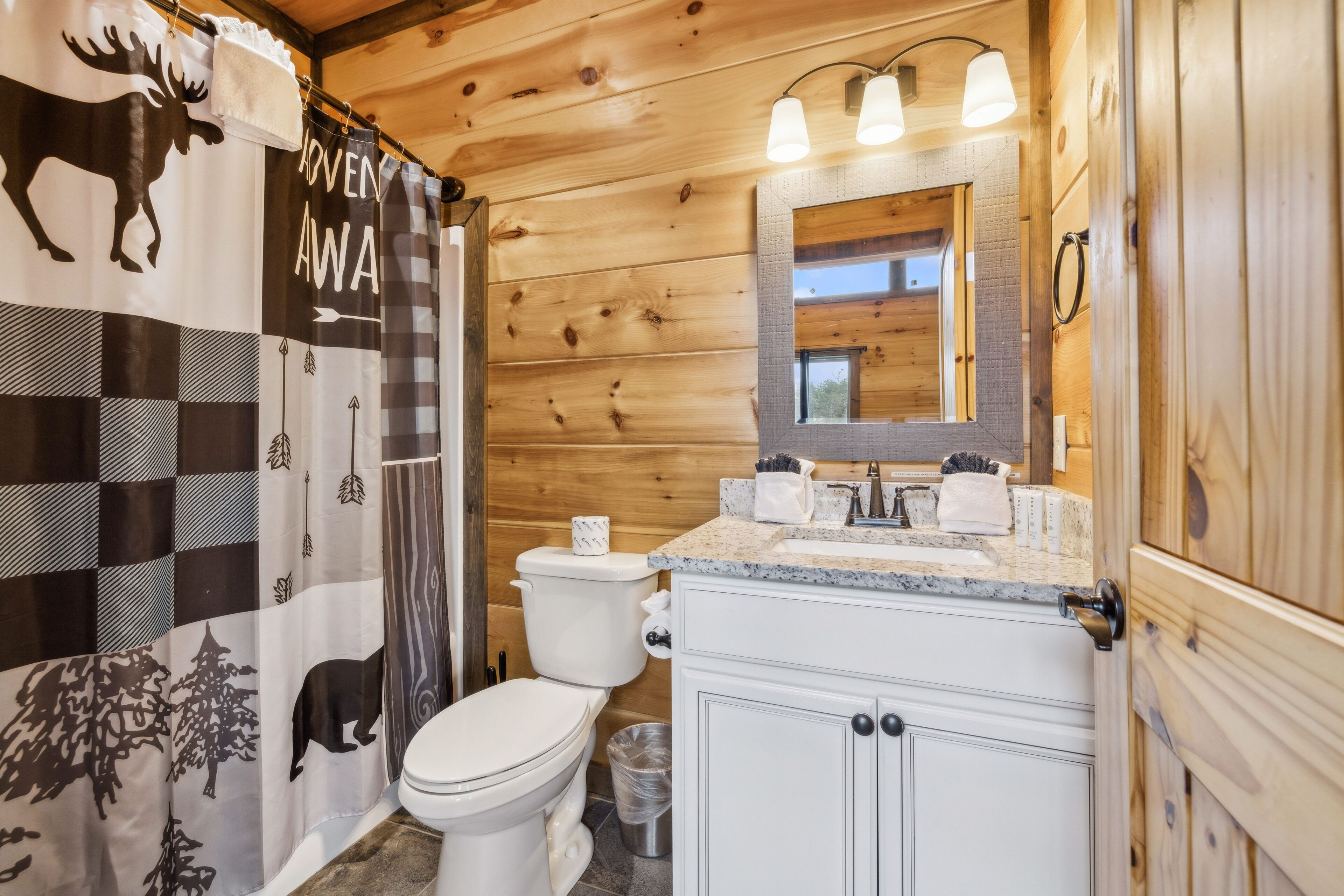 A wooden cabin bathroom with a checkered shower curtain, a toilet, a sink with a mirror, and a trash can.