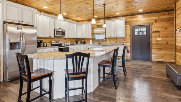 A kitchen with wooden cabinets, a stainless steel refrigerator, a microwave, and a large island with bar stools. - Eden Of The Smokies 5 Bedroom Cabin (Sevierville)