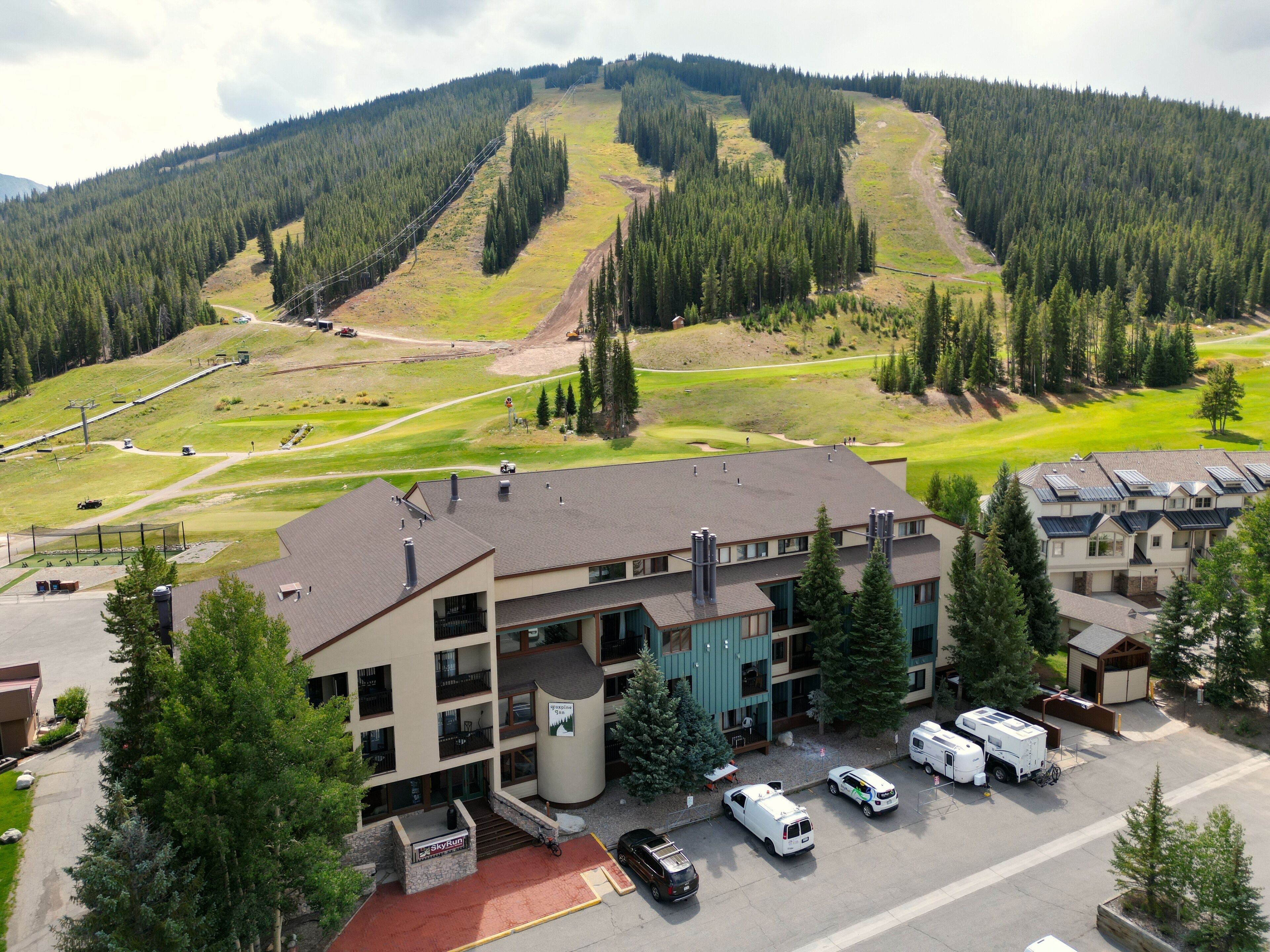 A multi-story hotel with a ski lift in the background, parking area, and a view of a mountainous landscape.