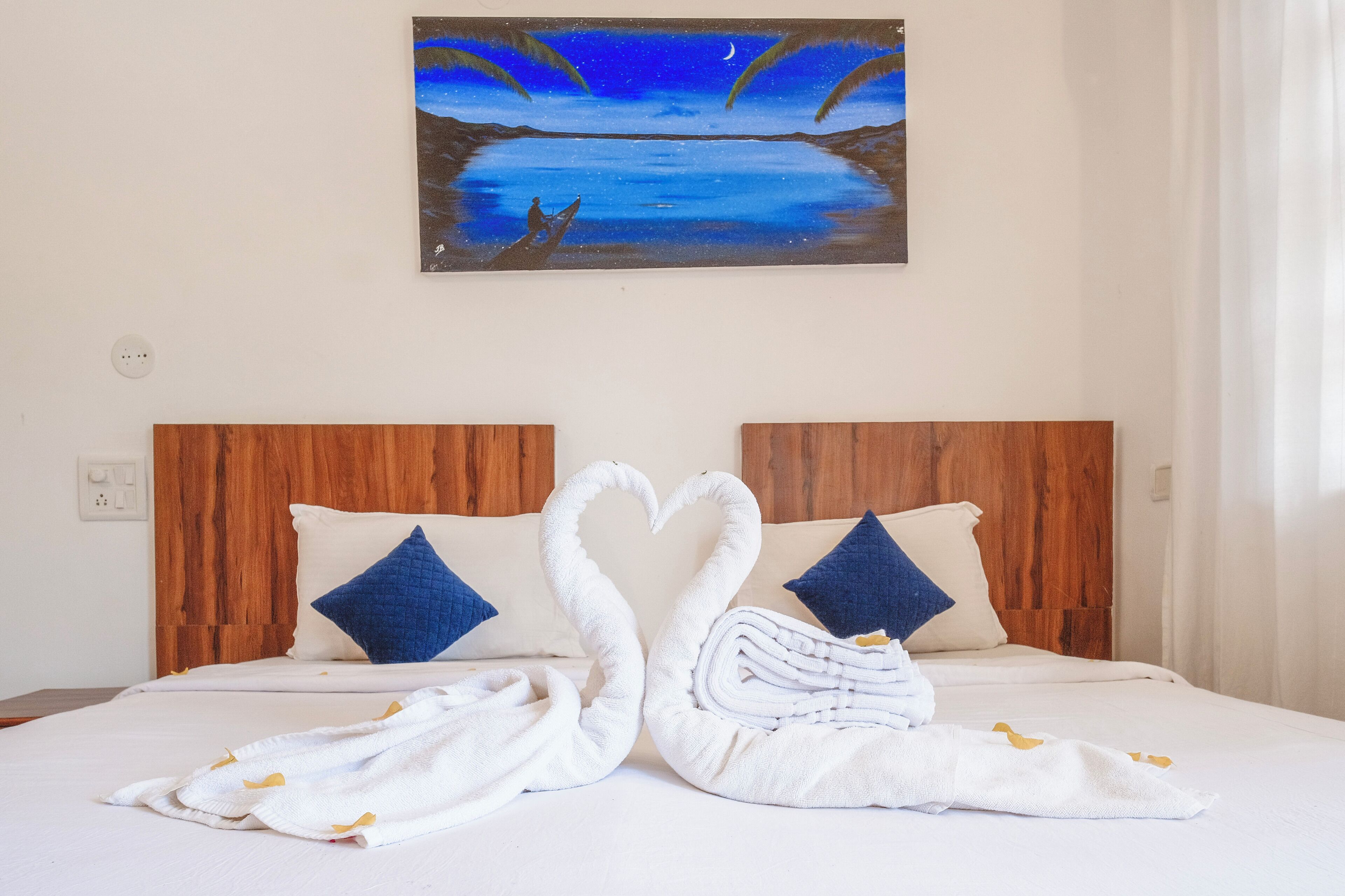 A hotel room with a bed featuring a swan-shaped towel arrangement, two blue pillows, and a painting of a night scene above the bed.