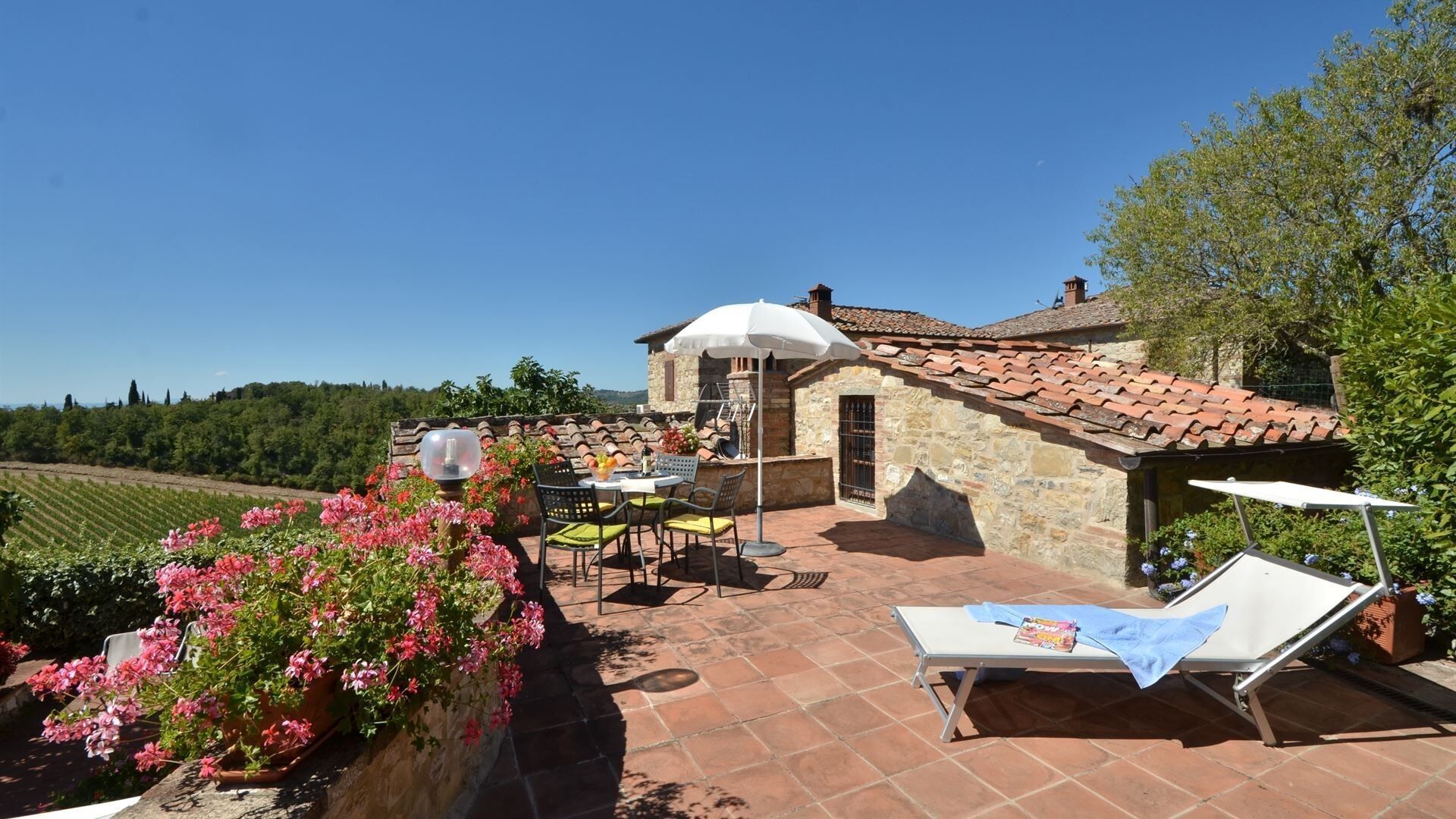 Un bâtiment en pierre avec un toit en tuiles, une terrasse avec une table et des chaises, et un jardin agrémenté de fleurs roses.