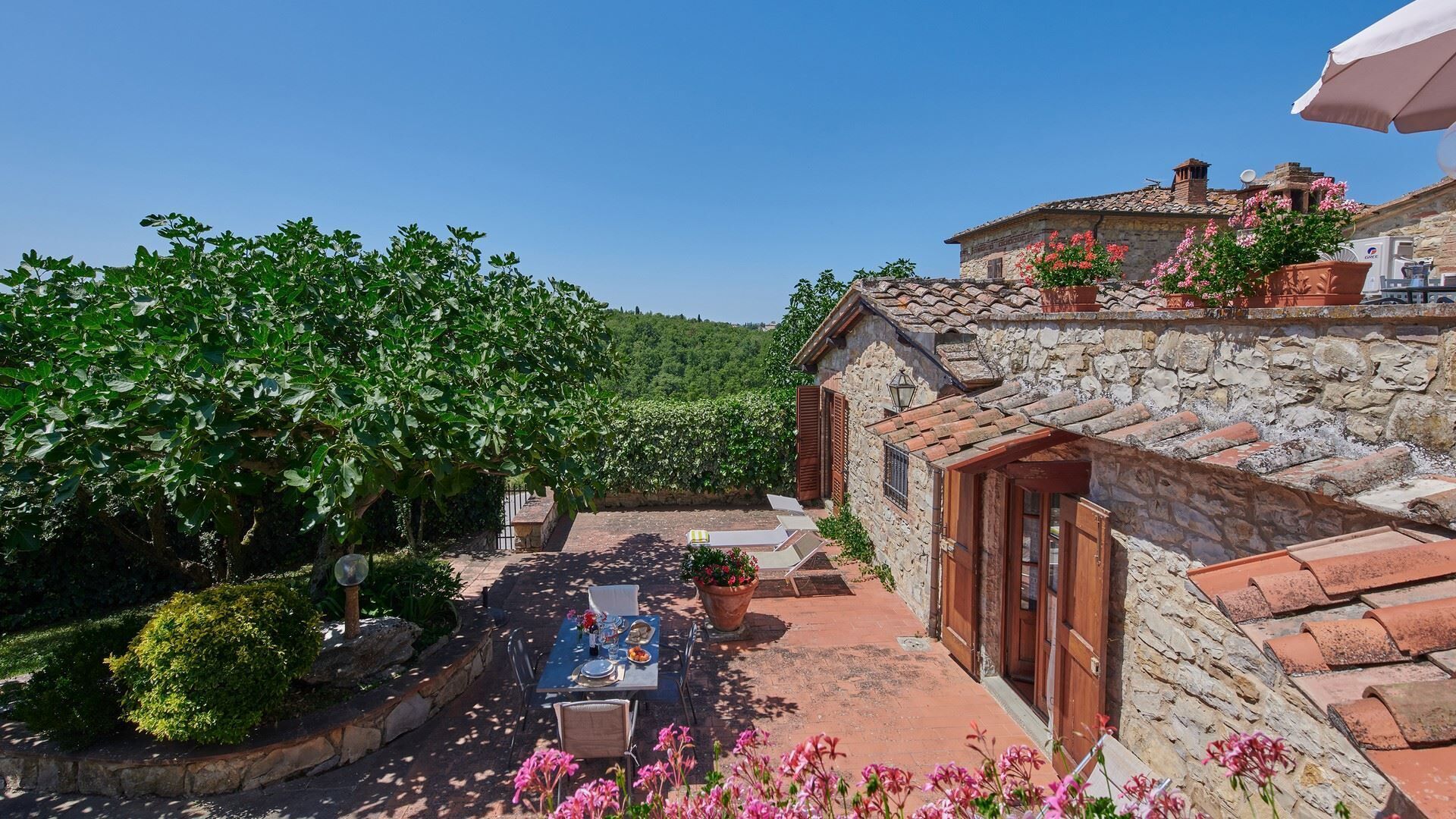 Un bâtiment en pierre avec un toit en tuiles, un jardin avec une table et des chaises, et une vue sur un vignoble.