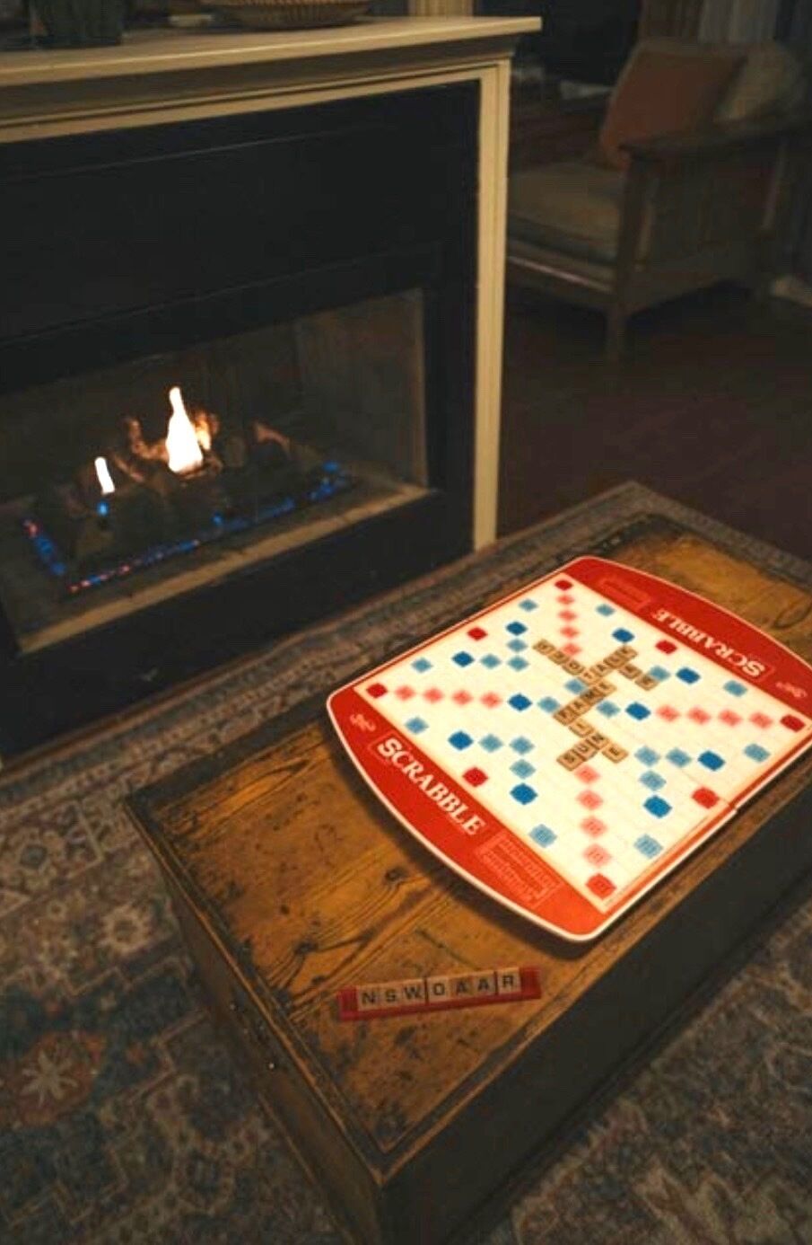 A Scrabble game on a wooden table with a fireplace in the background.