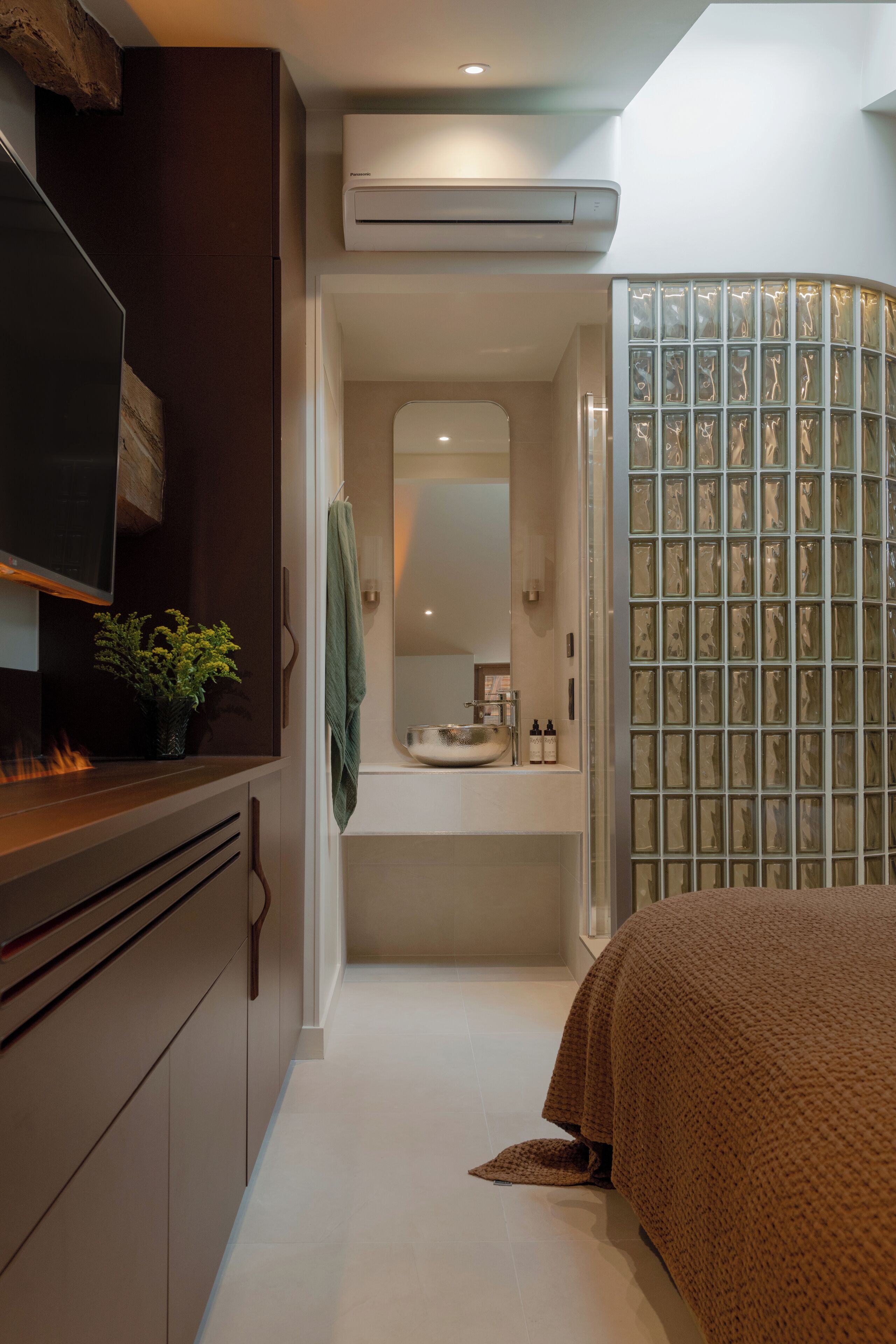 Una habitación de hotel estrecha con un armario de madera, televisión, una planta pequeña, y un baño con espejo, lavabo y cabina de ducha de vidrio.