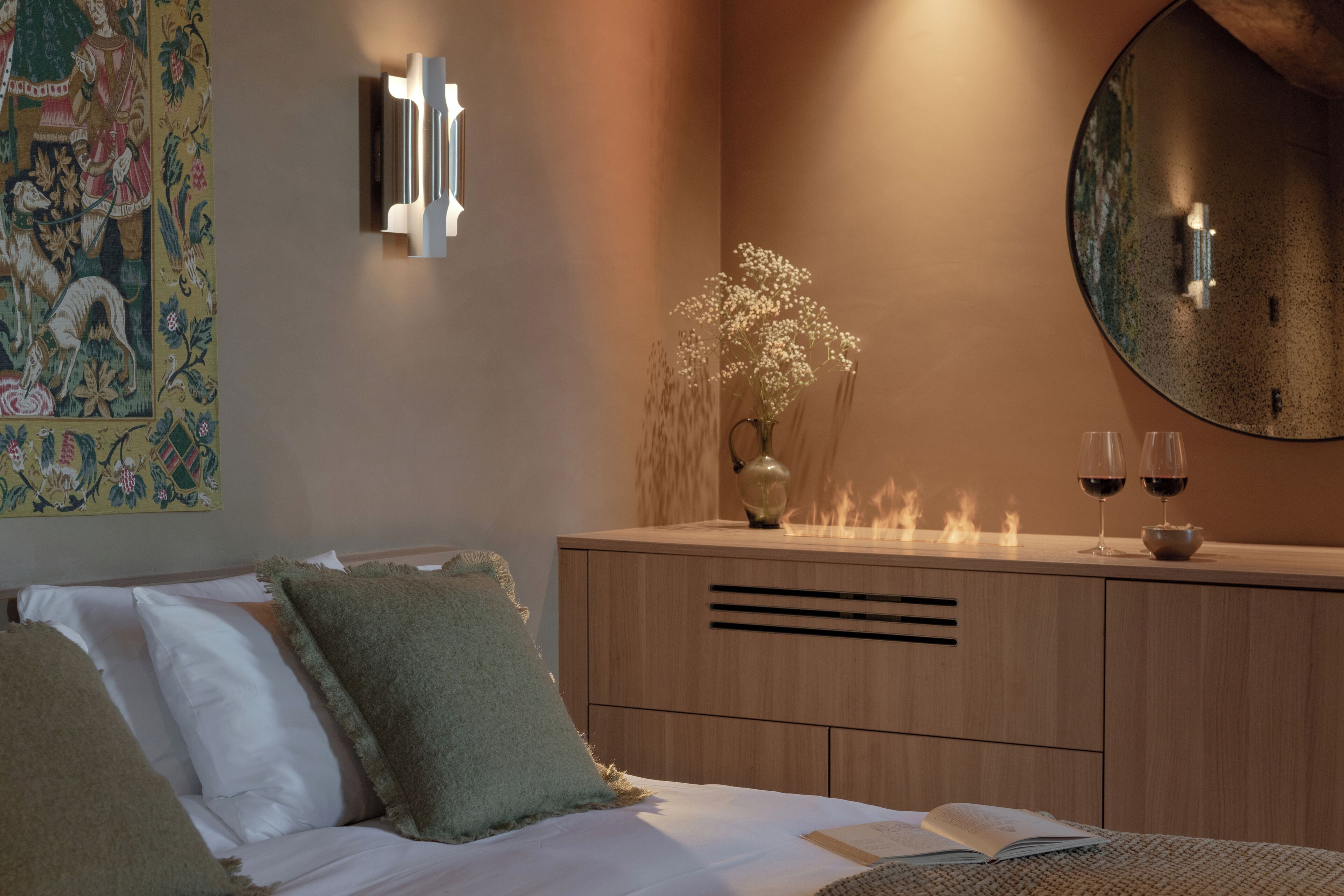 Ein Schlafzimmer mit einem Bett, Kissen, einem Nachttisch mit einer Vase und zwei Weingläsern, einem wandmontierten Spiegel und einem Wandteppich.