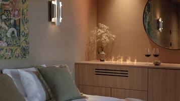 Ein Schlafzimmer mit einem Bett, Kissen, einem Nachttisch mit einer Vase und zwei Weingläsern, einem wandmontierten Spiegel und einem Wandteppich.