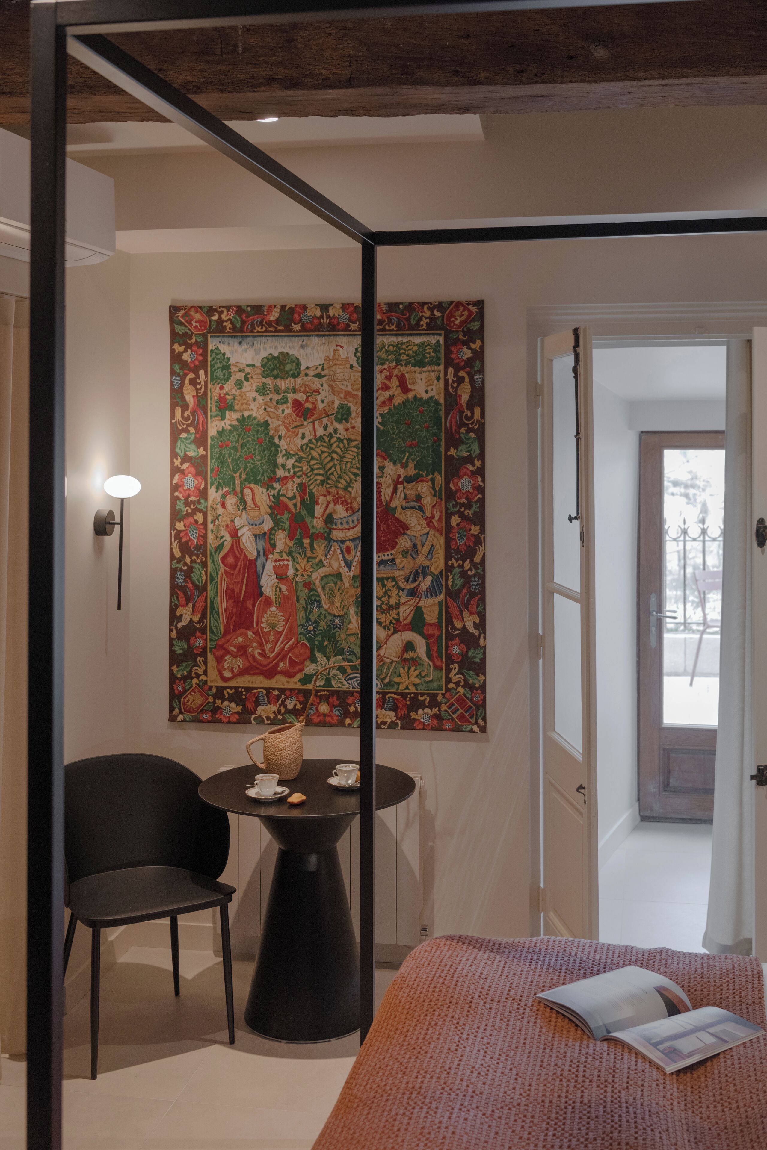 Une chambre à coucher avec une grande tapisserie, une petite table ronde avec une chaise, et un lit sur lequel se trouve un magazine.
