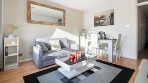 A modern living room with a grey sofa, a glass coffee table, and a dining area with white chairs.