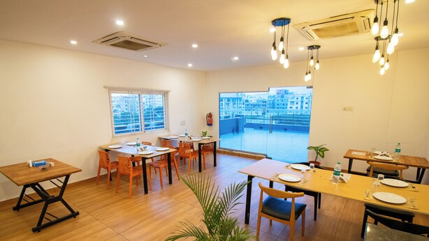 A dining area with multiple tables and chairs, a large window with blinds, and a view of buildings outside. - Moonlight Inn (Bengaluru)