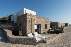 A stone building with a white upper section, a small patio with white lounge chairs, and a table. - Ocean Stone Suite: Overlooking the Aegean Sea! (Παροικιά)