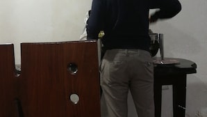 A person standing in a room with a wooden table and a black chair. There is a white wall with a power outlet and a switchboard. - Eco mara forest camp  (Meru)