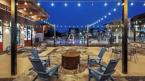 An outdoor patio area with seating and a fire pit, surrounded by string lights and modern buildings in the background. - Boulevard Stays Direct Trail Access! Parking, Pool, Gym 04 (RICHARDSON)