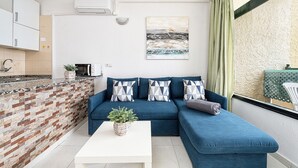 Un espacio habitable compacto con área de cocina, un sofá azul con cojines estampados, una mesa de centro blanca y una planta en maceta.