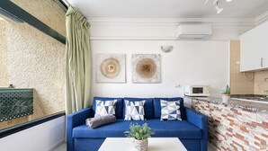 Un salón con un sofá azul, una mesa de centro blanca y una cortina verde. Hay dos obras de arte en la pared sobre el sofá.