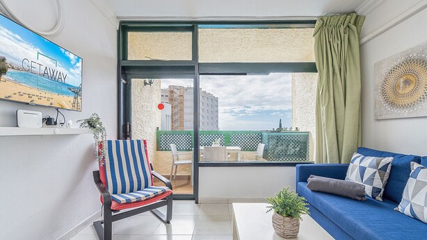 Un balcón con vista a la playa, un sofá azul, una silla a rayas y una pequeña mesa con una planta.