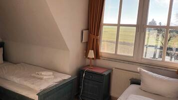 A bedroom with a single bed, a nightstand, a lamp, a window with a view of the countryside, and a radiator.