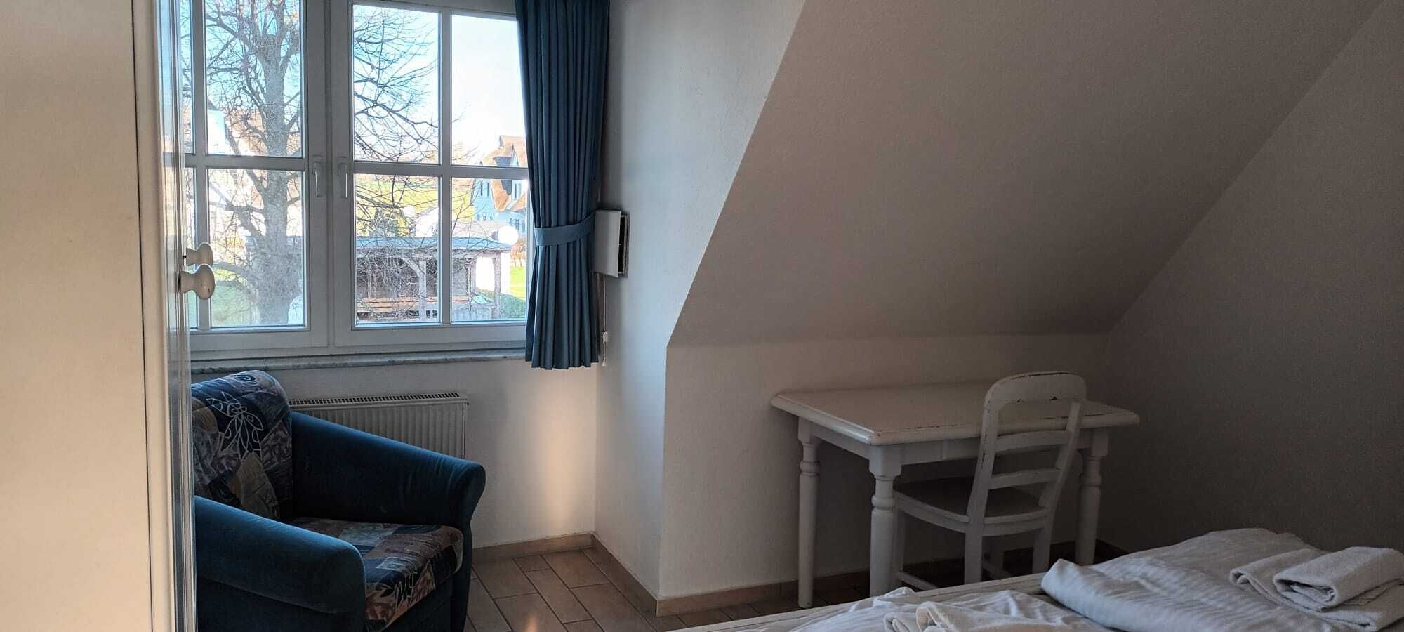 Ein kleiner, gemütlicher Raum mit schrägen Wänden, einem Bett, einem Sessel, einem Tisch und einem fenster mit Vorhängen.