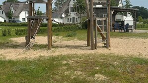A wooden play structure in a grassy area with houses in the background. - Landhaus am Haff, Apartment B6 - Vacation under thatch (Stolpe auf Usedom)