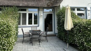 A house with a thatched roof, a patio with a table and chairs, and a large umbrella. - Landhaus am Haff, Apartment B6 - Vacation under thatch (Stolpe auf Usedom)