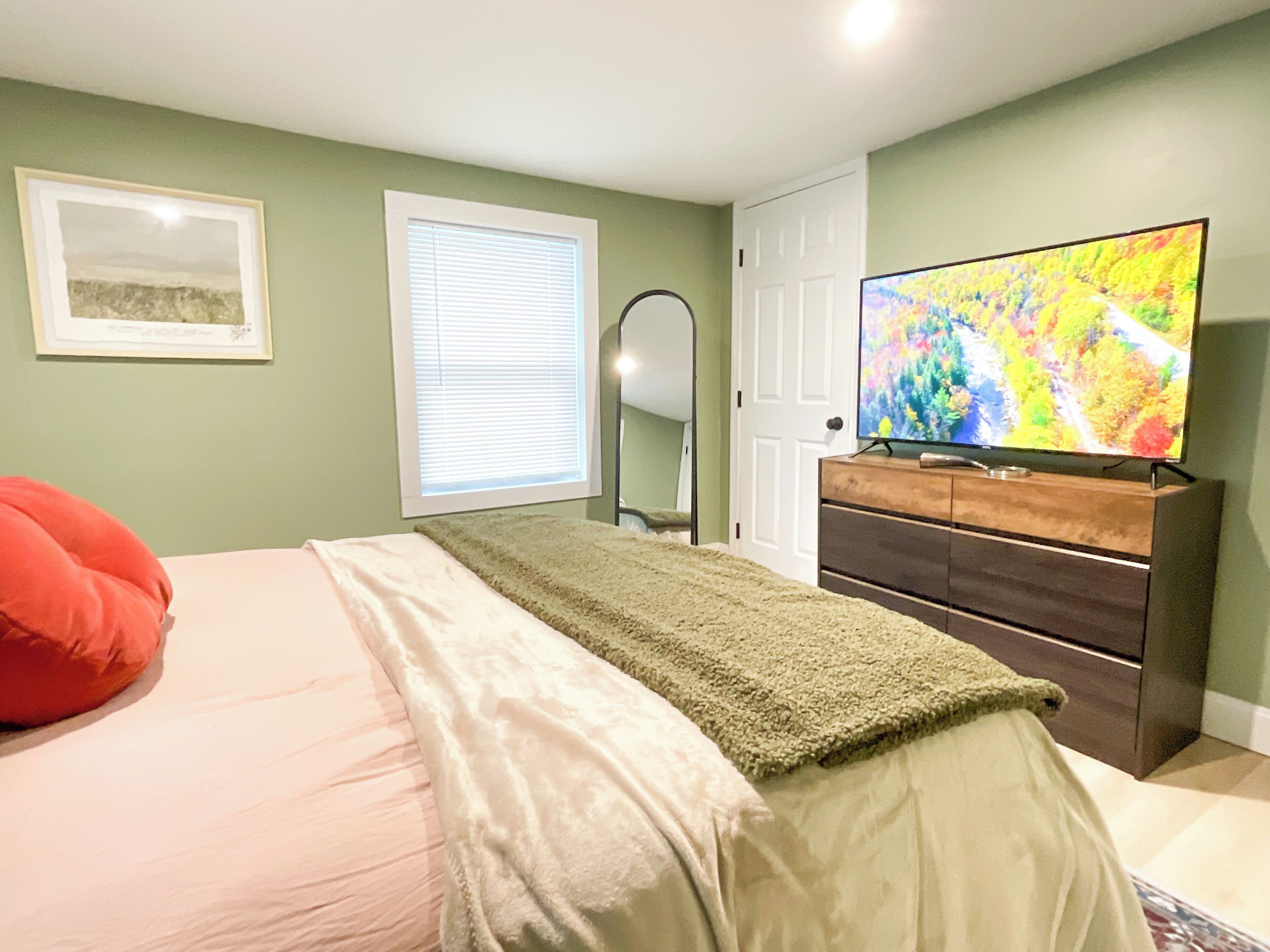 A bedroom with a bed, a dresser, a television, a window with blinds, and a door.
