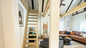 Una escalera conduce a una zona de altillo con una escalera de madera, un sofá gris y una mesa de centro en una sala de estar con alfombra estampada.