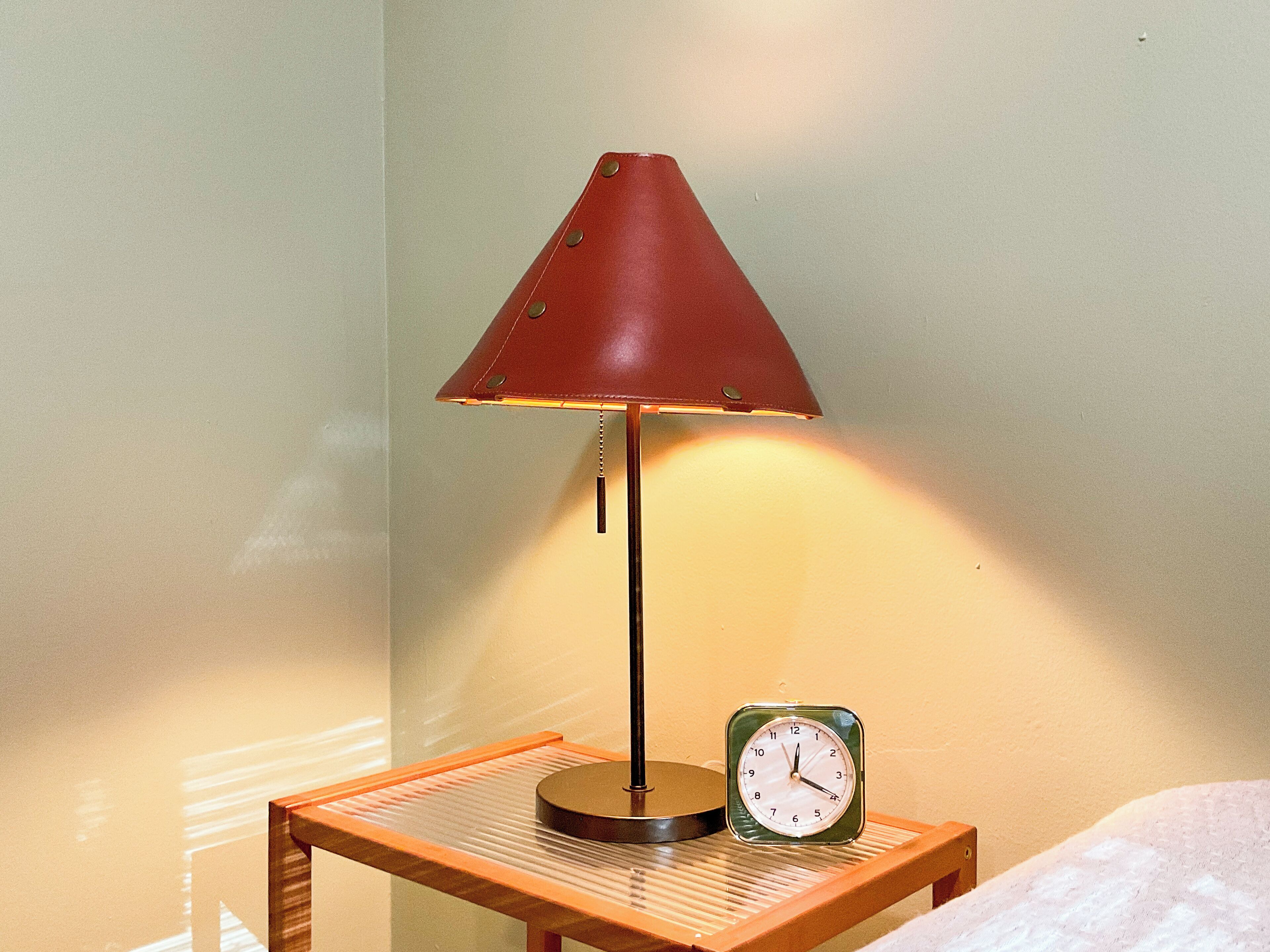 Eine Lampe mit kegelförmigem Lampenschirm und eine Uhr auf einem Holztisch vor einer Wand.