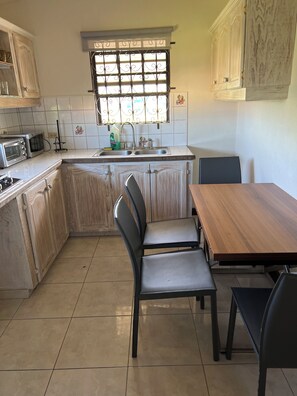 A kitchen with a dining area, featuring a wooden table, two chairs, a sink, and a window with decorative iron bars. - Cozy place with WiFi, AC ,close to beach. Free loaner car avail. (Saint James)
