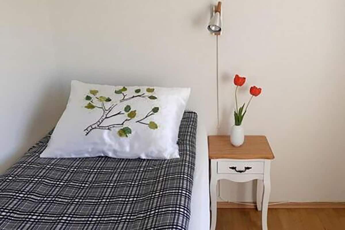 Un lit avec un oreiller blanc orné d’un motif de branches et de feuilles, une couvre-lit à carreaux noirs et blancs, et une table de chevet sur laquelle se trouve un vase blanc contenant des fleurs rouges.