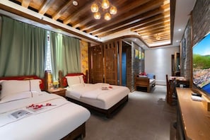 A hotel room with two beds, wooden ceiling beams, a stone wall, and a view of greenery.