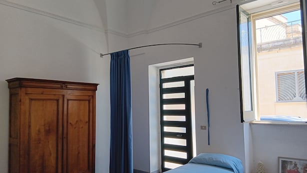 A room with a bed, a wooden wardrobe, a door with a curtain, and a window with a view of the outside. - Casetta Rita in Melpignano (Melpignano)