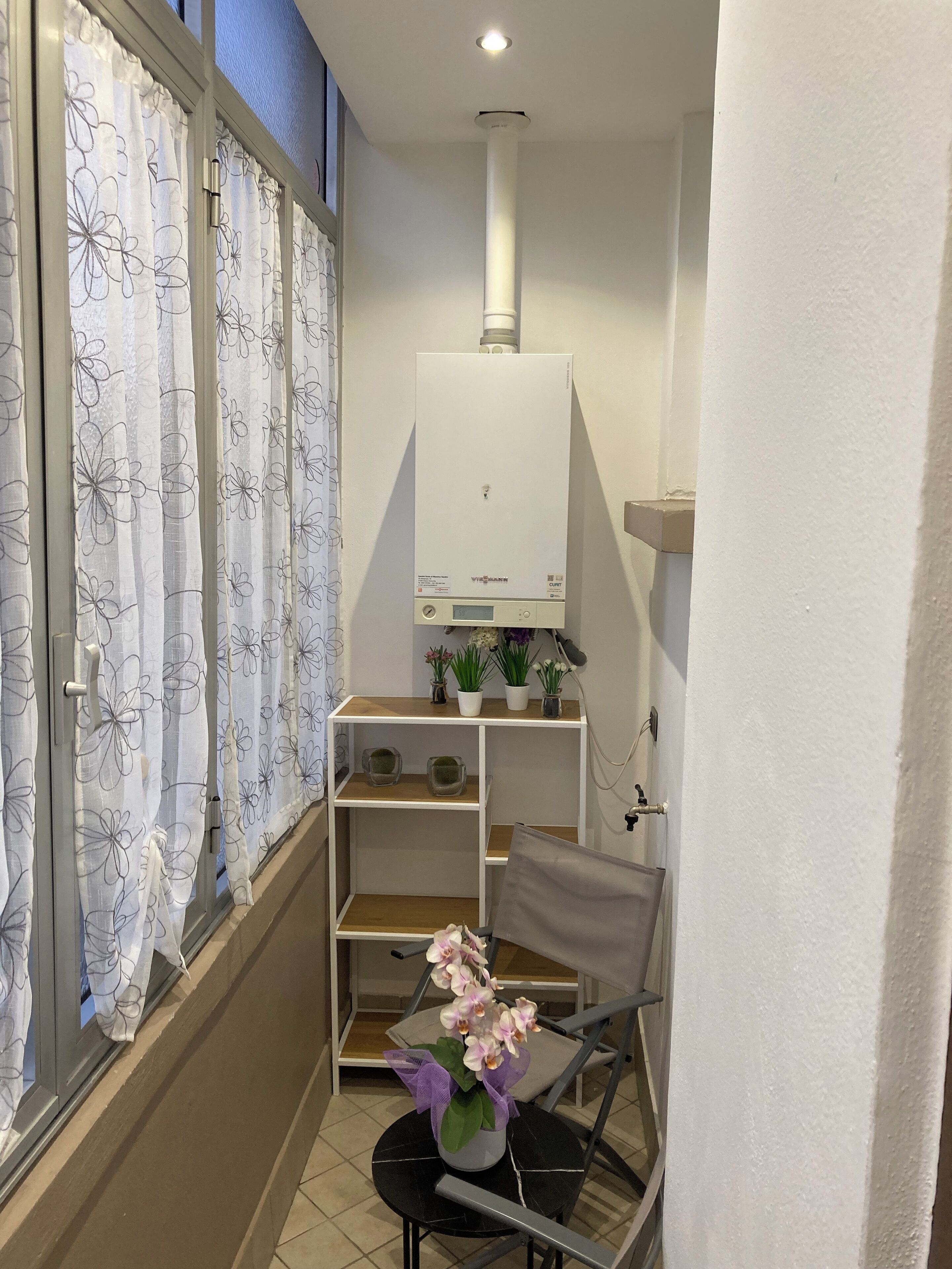 Une petite pièce étroite, avec un radiateur, une chaise, une petite table et une fenêtre ornée de rideaux à motifs floraux.