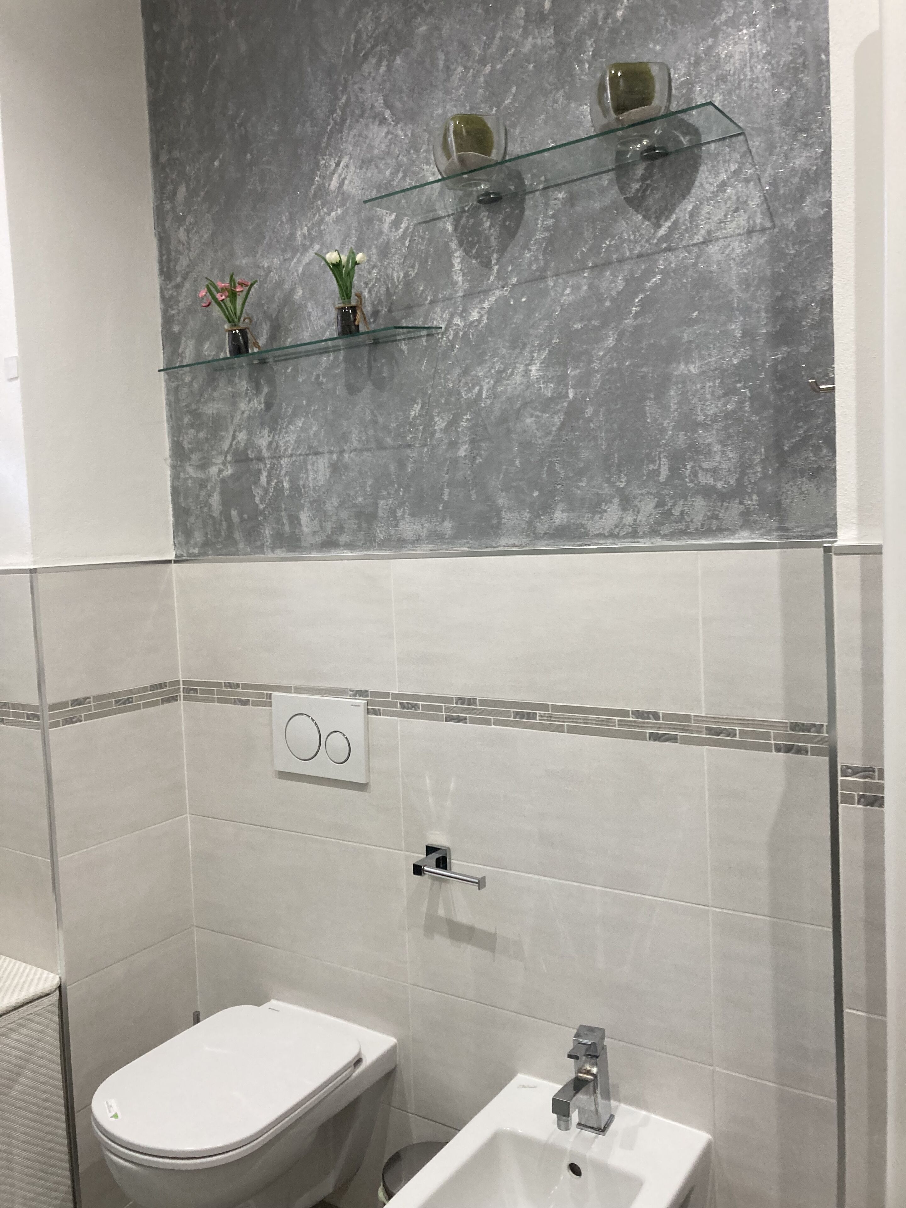 Un baño con inodoro, lavamanos y repisas de vidrio frente a una pared con textura.