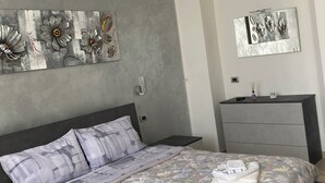 A modern bedroom with a grey and white color scheme, a bed with floral bedding, a grey dresser, and two wall art pieces. - Orchid Apartment (Castiglione Olona)