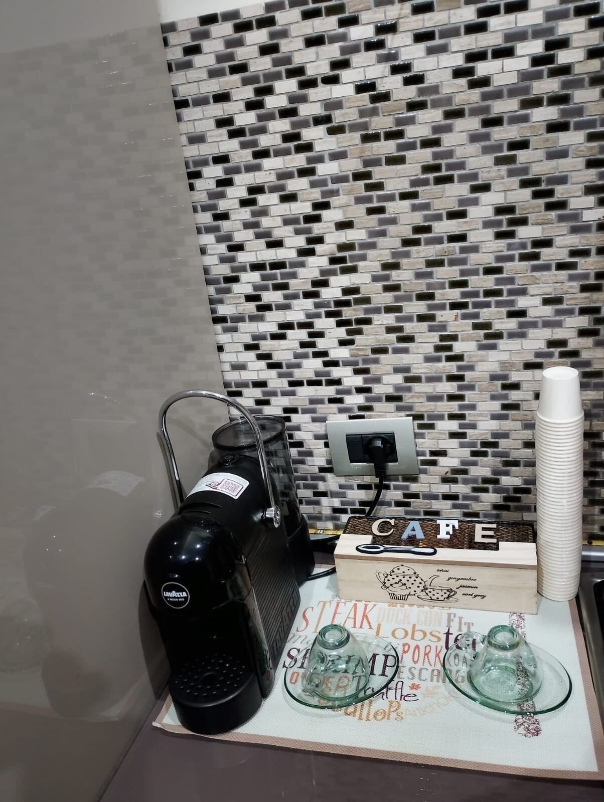 Un coin cuisine équipé d’une machine à café, d’une boîte étiquetée « CAFÉ » et d’un dosseret carrelé.