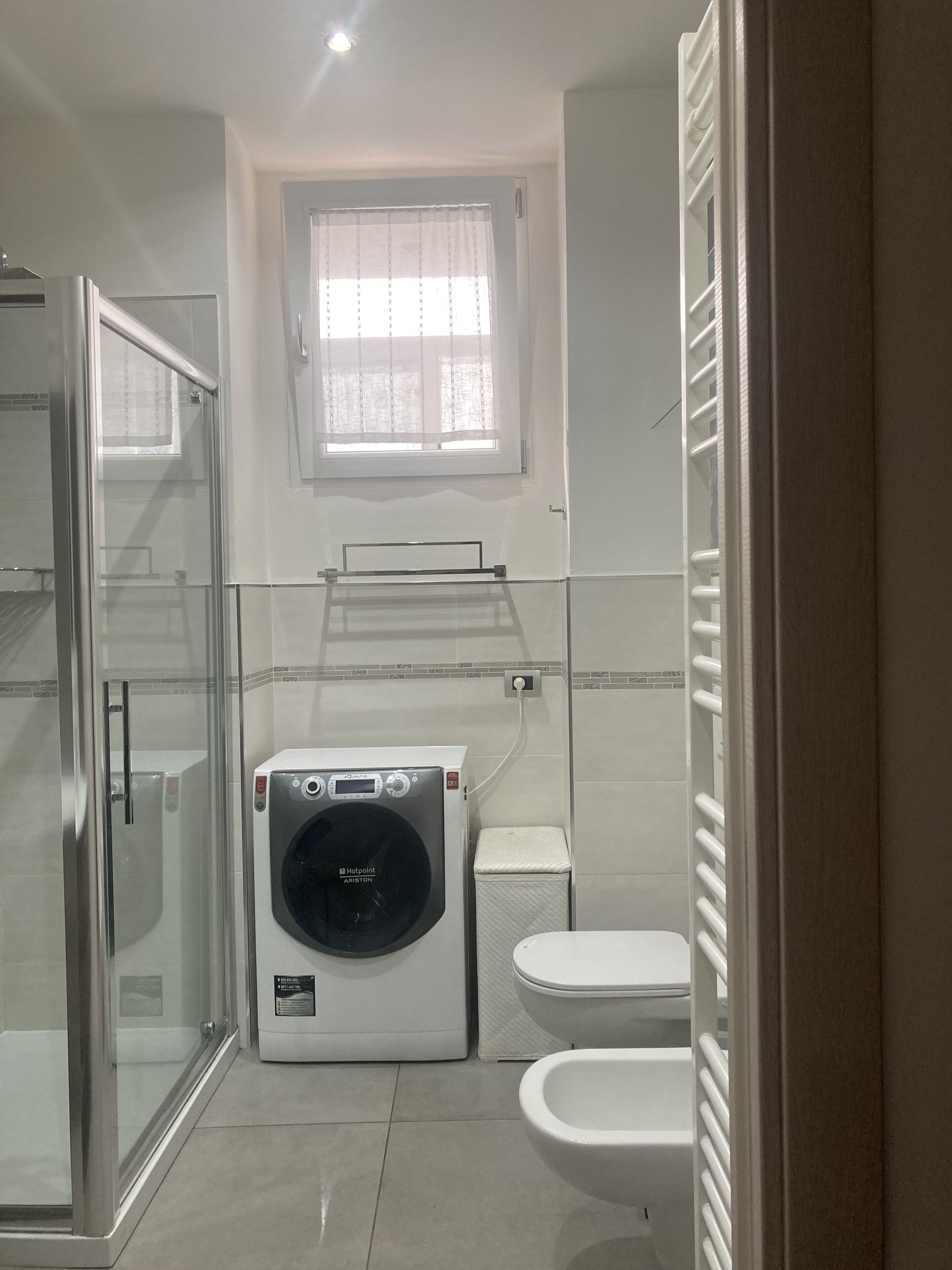 Ein kleines Badezimmer mit Duschkabine aus Glas, Waschmaschine, Toilette und Fenster mit leichtem Vorhang.