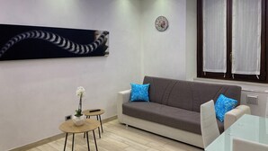 A modern living room with a grey sofa, two blue pillows, a round table with a vase of white flowers, a clock on the wall, and a window with curtains. - Orchid Apartment (Castiglione Olona)