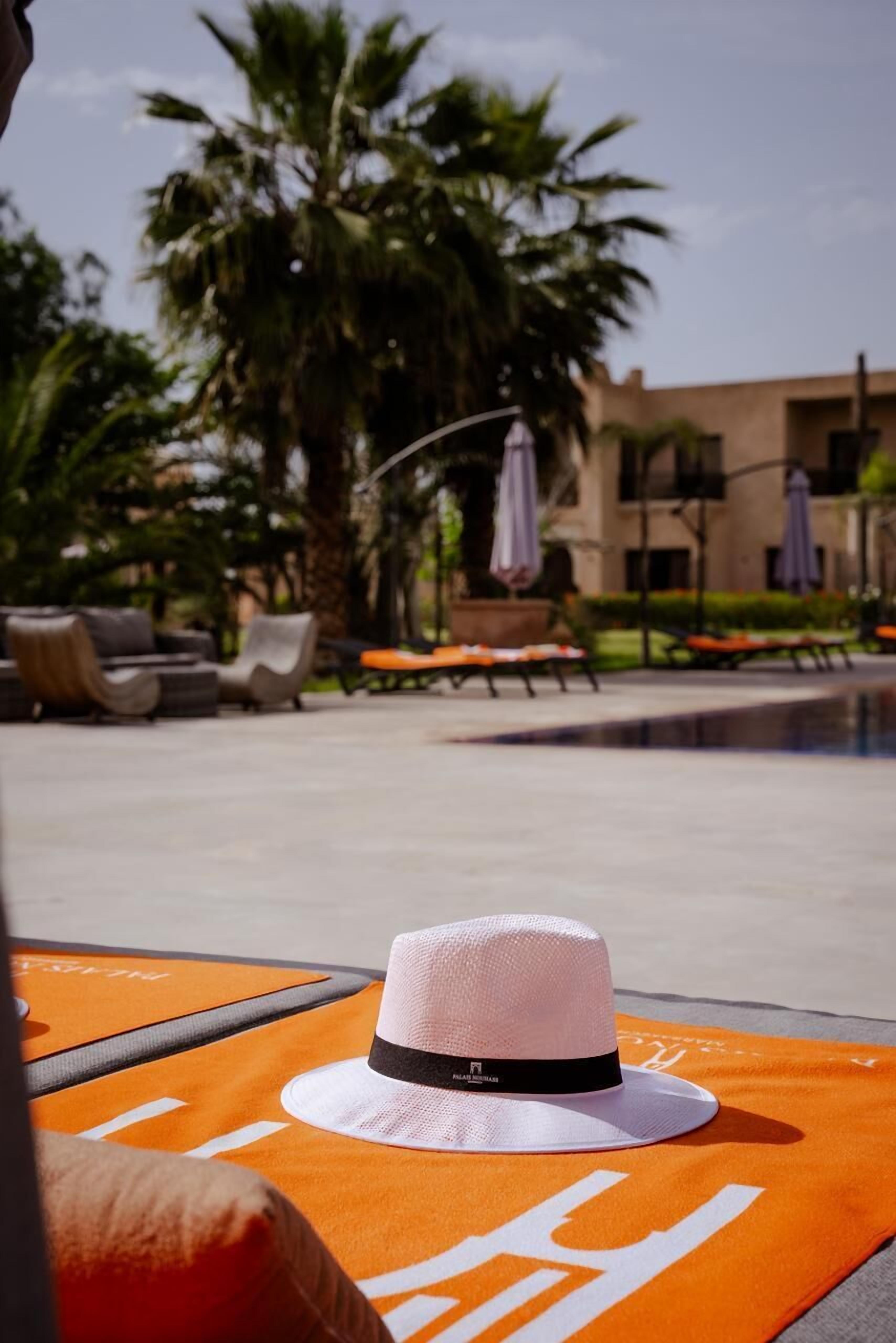 Un espace aménagé au bord de la piscine, avec des chaises longues, un chapeau posé sur une serviette et des palmiers en arrière-plan.