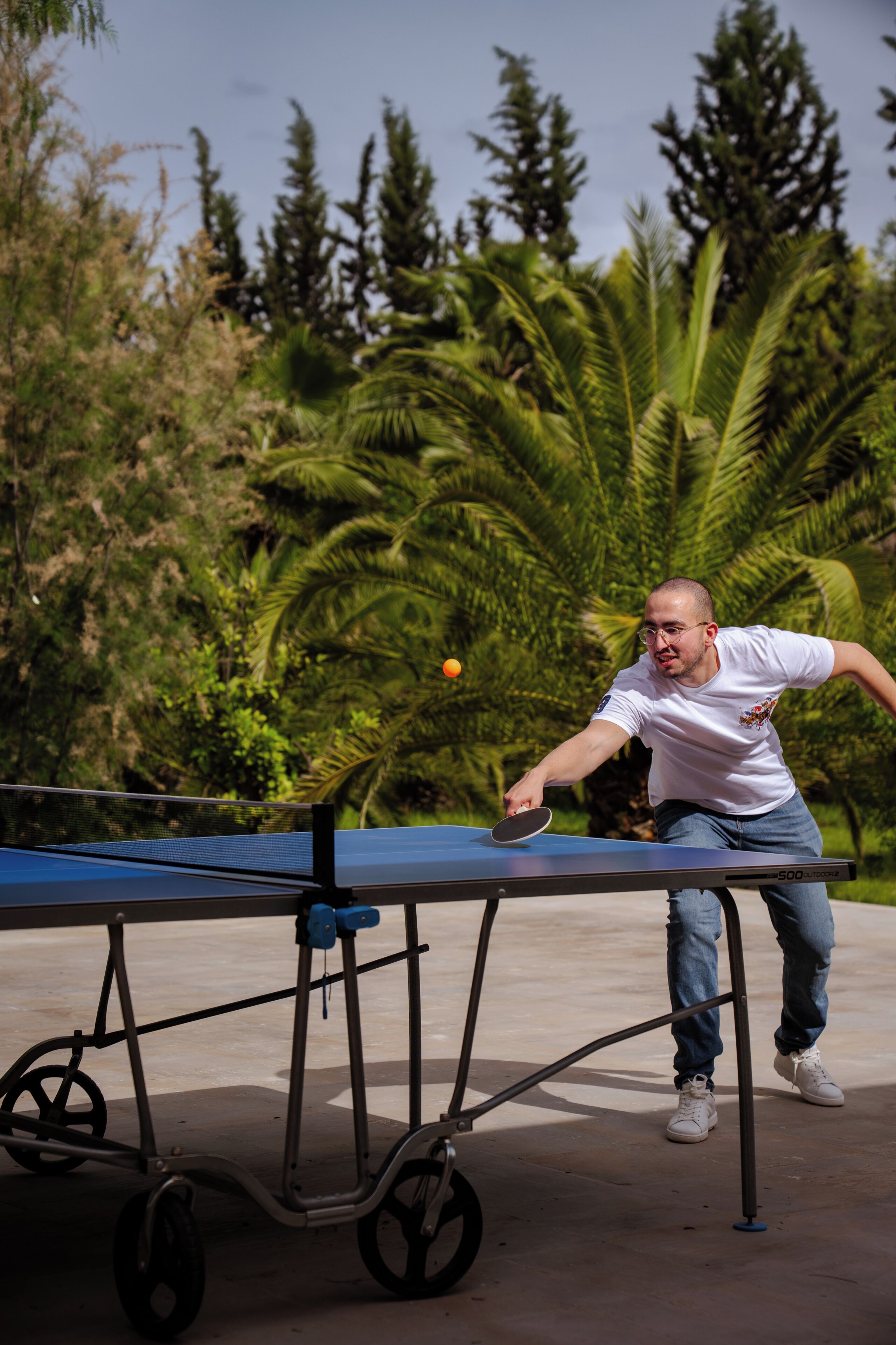 A man playing table tennis outdoors with a table on wheels, surrounded by greenery.