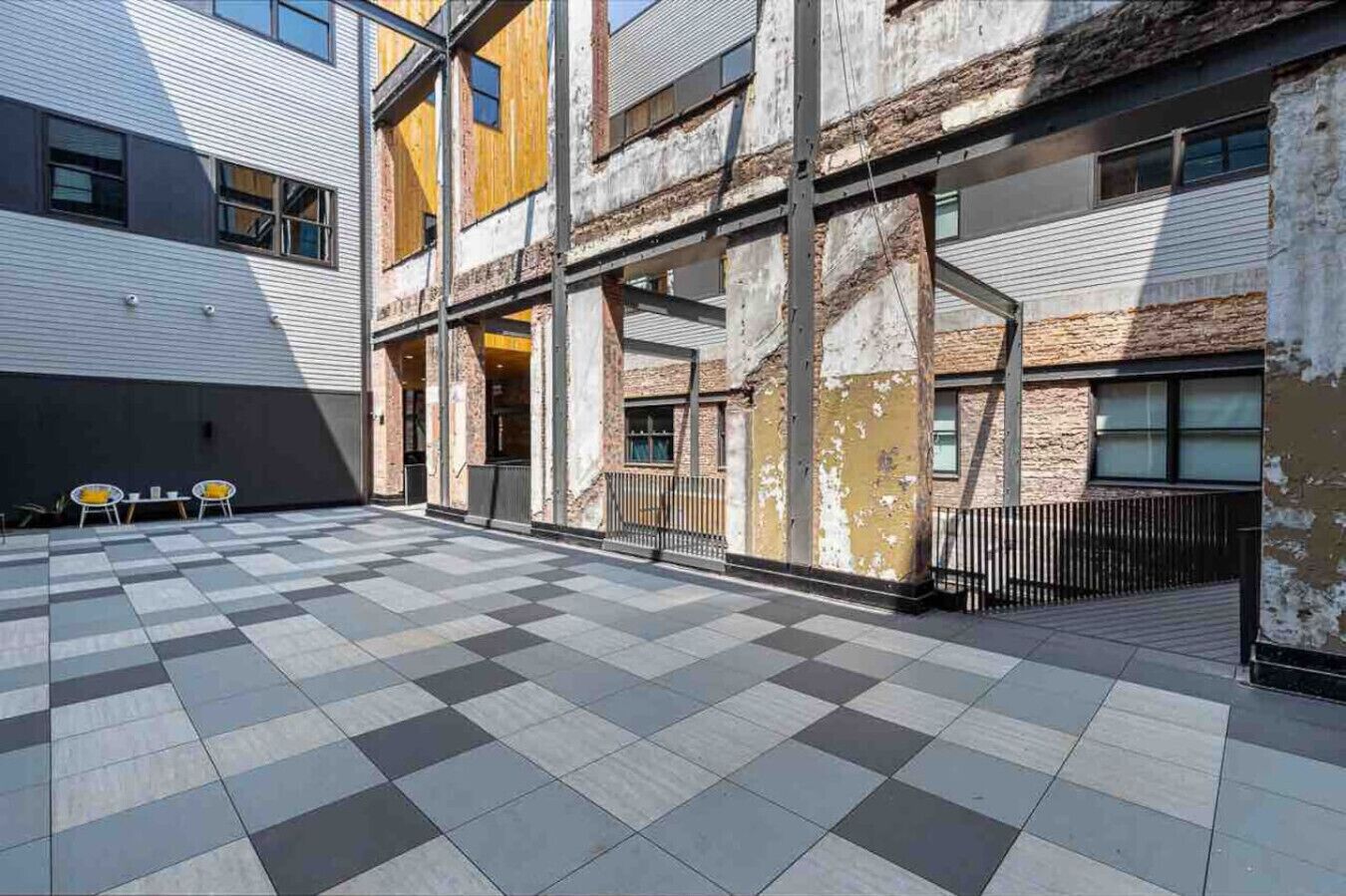 An outdoor courtyard with a checkered floor, metal railings, and a building with exposed brick and wooden elements.