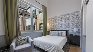 A bedroom with a bed, a chair, a nightstand, and a window with curtains. - #302 New Downtown Condo on Morris Ave (Birmingham)