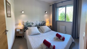 A neatly made bed with two pillows and two red towels, a nightstand with a lamp, a framed picture on the wall, and a window with curtains. - Waterfront apartment, Porto-Vecchio (Porto-Vecchio)