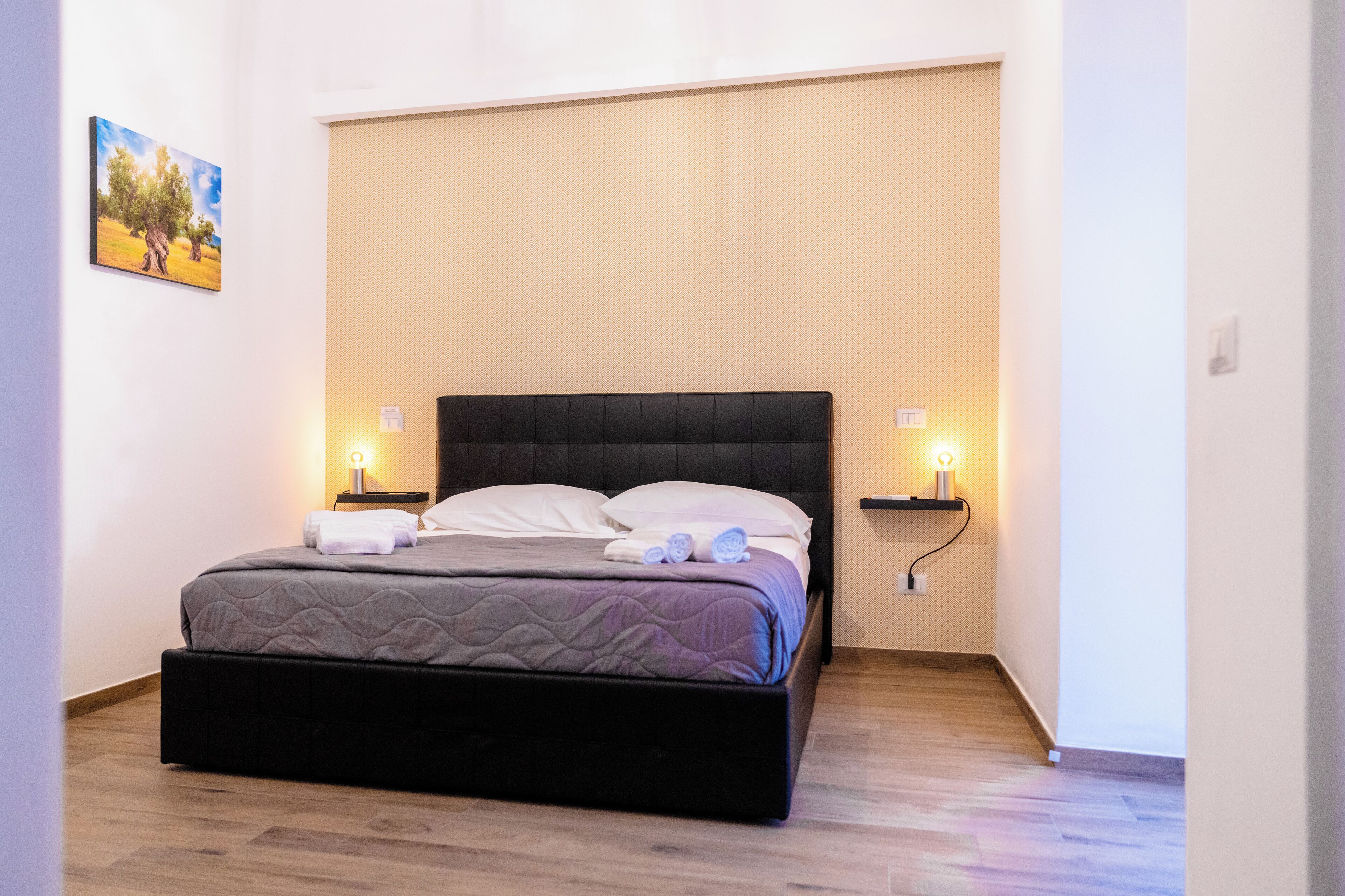 Una cama bien tendida con un cabecero oscuro, dos mesitas de noche con lámparas y un cuadro colgado en la pared.