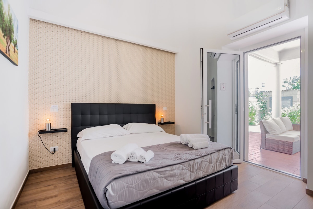 A modern bedroom with a large bed, a nightstand with a lamp, and a sliding glass door leading to an outdoor seating area.