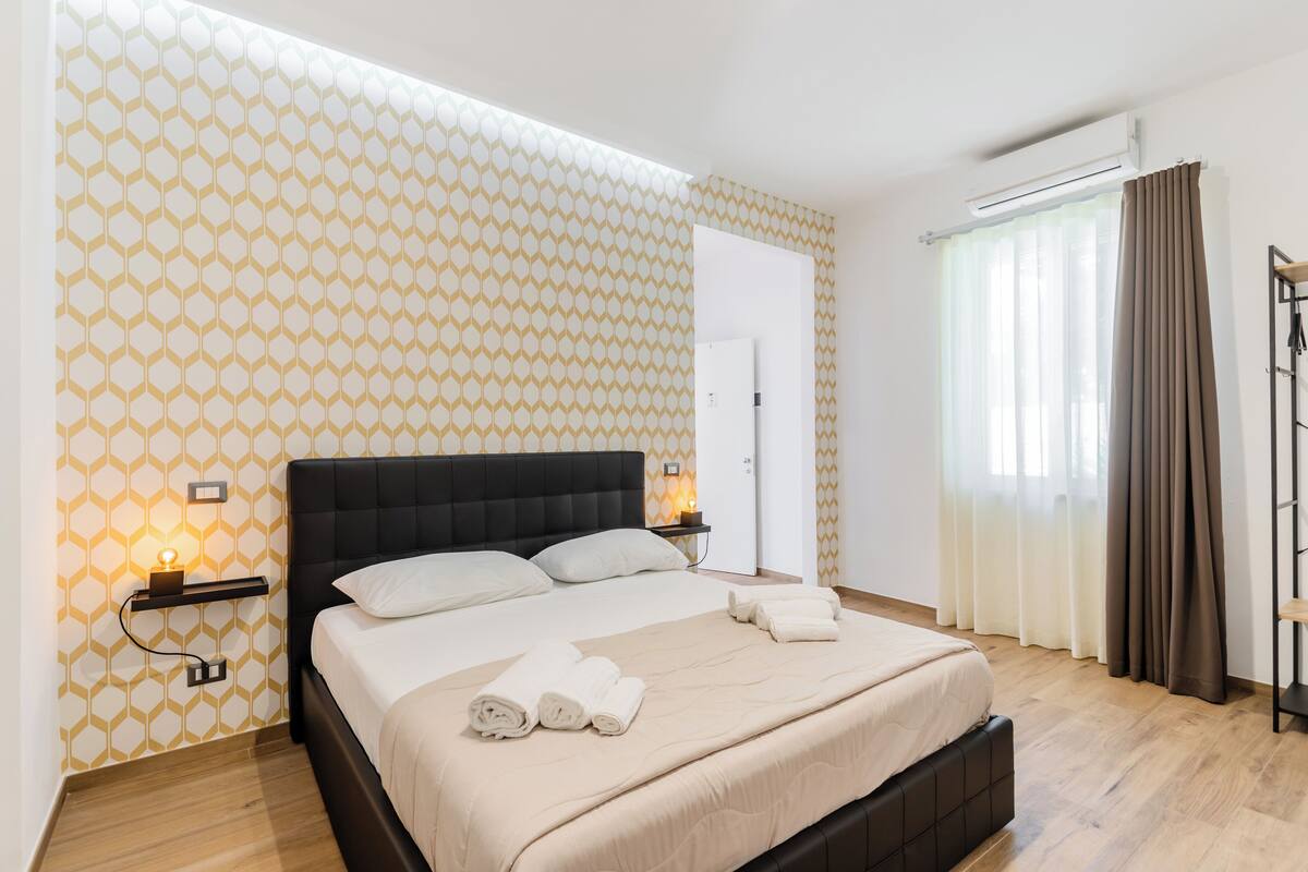 A modern bedroom with a geometric patterned wall, a black headboard, a bedside table with a lamp, and a neatly made bed with towels.
