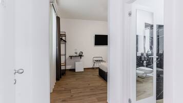 A modern hotel room with a wooden floor, a bed, a TV mounted on the wall, and a bathroom visible through a glass door.
