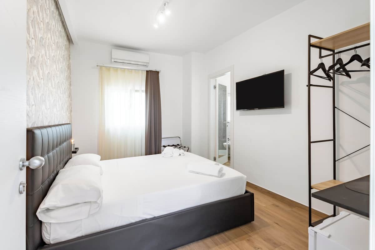 A modern hotel room with a bed, a TV mounted on the wall, a wardrobe with hanging clothes, and an air conditioning unit.