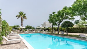 Una piscina con sillones de descanso y sombrillas, rodeada de vegetación y un edificio al fondo.