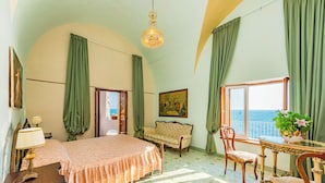 Een kamer met een bed, een slaapbank, een eettafel met stoelen, een kroonluchter en uitzicht op zee door een raam.