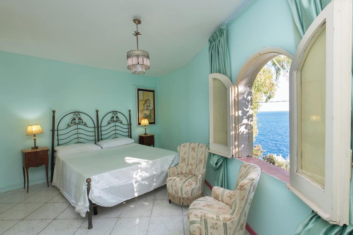 Un dormitorio con cama, dos mesitas de noche con lámparas, una silla, una ventana con vista al mar y una araña.