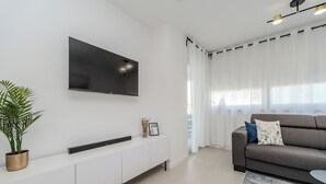 A modern living room with a flat-screen TV mounted on the wall, a white console table with decorative items, a grey sofa with cushions, and sheer curtains on a window. - Two-bedroom apartment with a terrace (Novalja)
